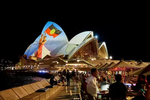 Photo of The Sydney Opera House with a woman waving an aboriginal flag proejcted onto the sails of the building.